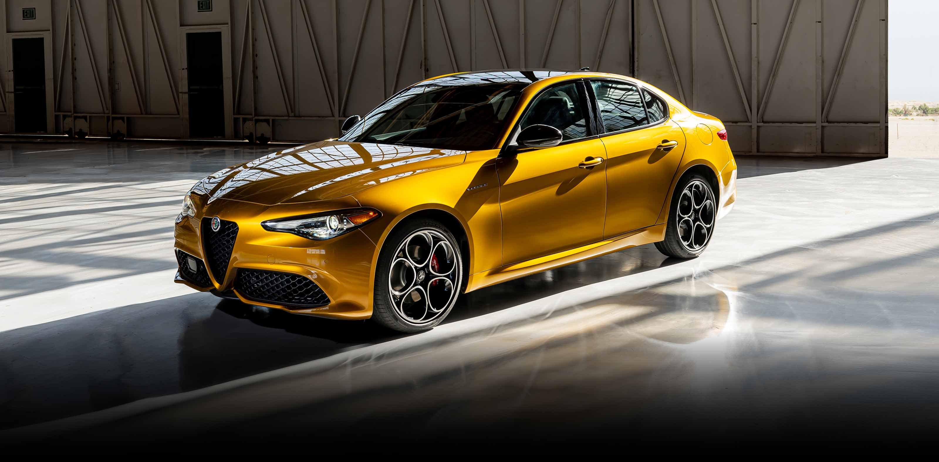 Un Alfa Romeo Giulia Veloce 2023 dorado estacionado en un garaje industrial, recibiendo luz proveniente de la puerta abierta del garaje.