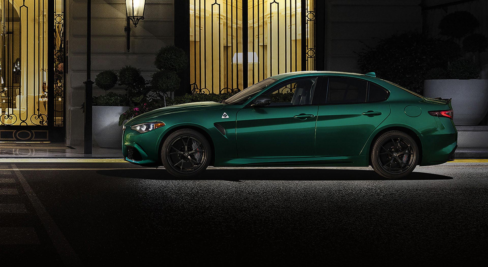 Perfil del lado del conductor de un Alfa Romeo Giulia Quadrifoglio 2024 verde estacionado frente a un edificio comercial por la noche.