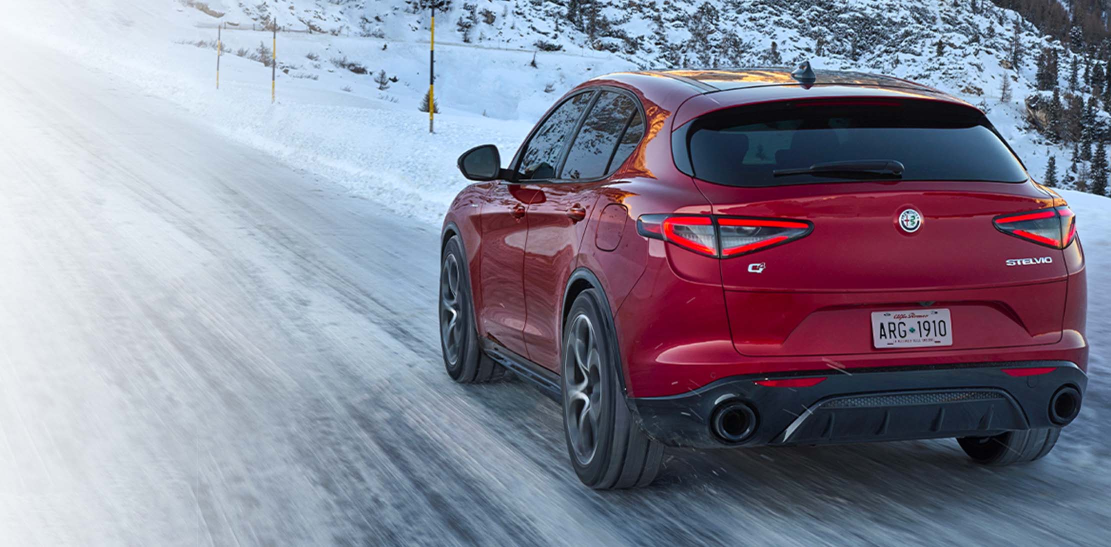 Ángulo trasero de un Alfa Romeo Stelvio TBD 2024 rojo circulando por un camino cubierto de nieve en las montañas.