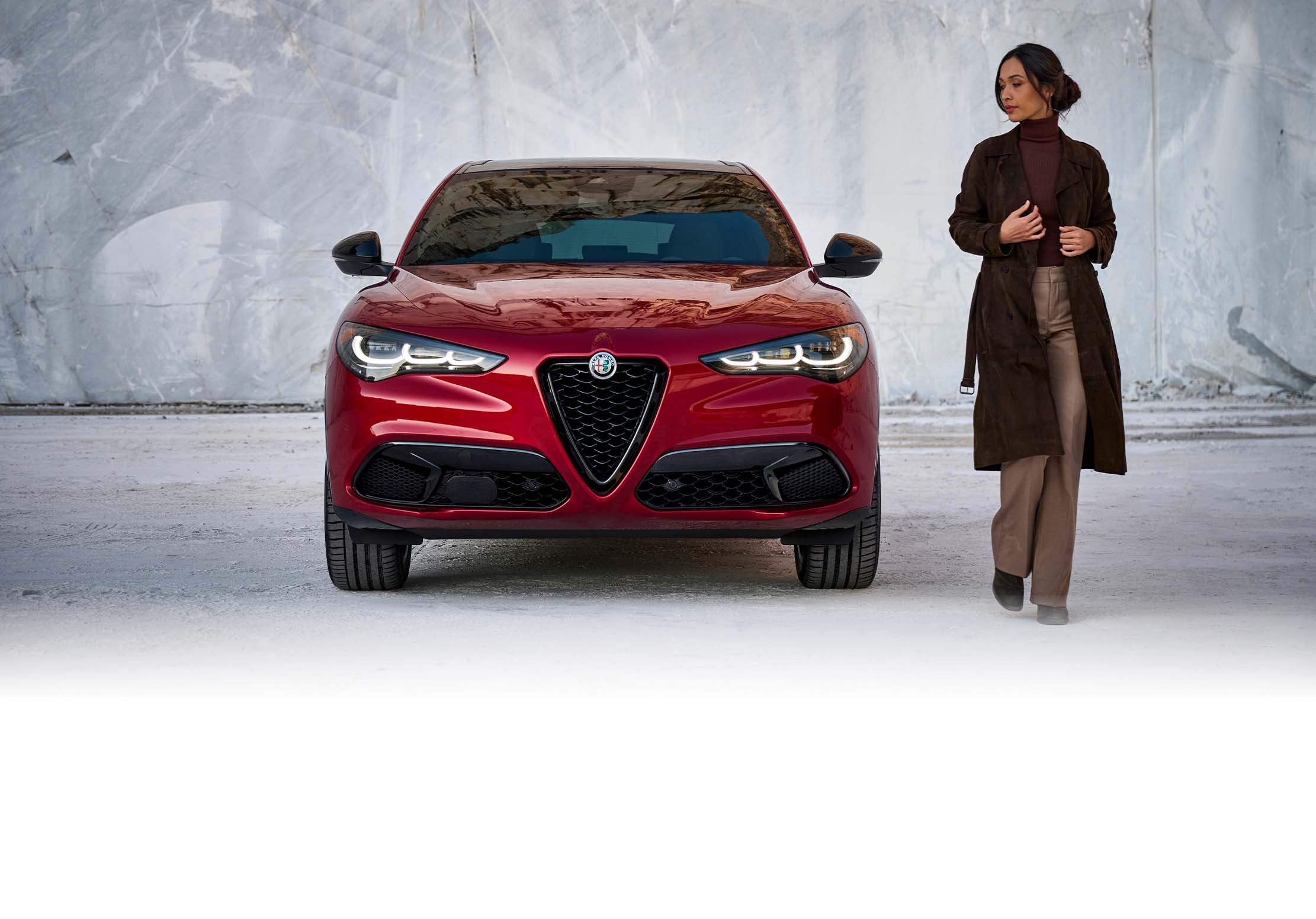 Un ángulo frontal del Alfa Romeo Stelvio TBD 2024 rojo estacionado frente a un muro de piedra mientras una mujer se aleja del vehículo.