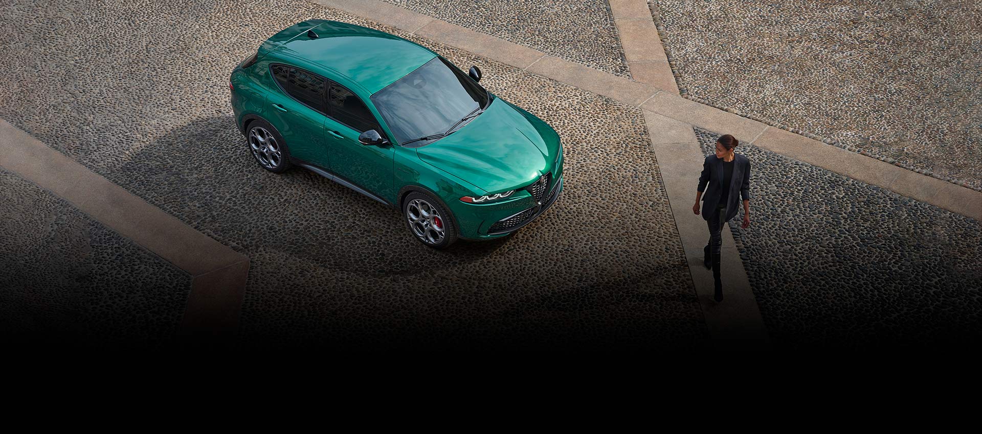 Una imagen desde un ángulo superior de un Alfa Romeo Tonale Veloce 2024 verde estacionado en un patio. Una mujer pasa junto al vehículo y lo mira.