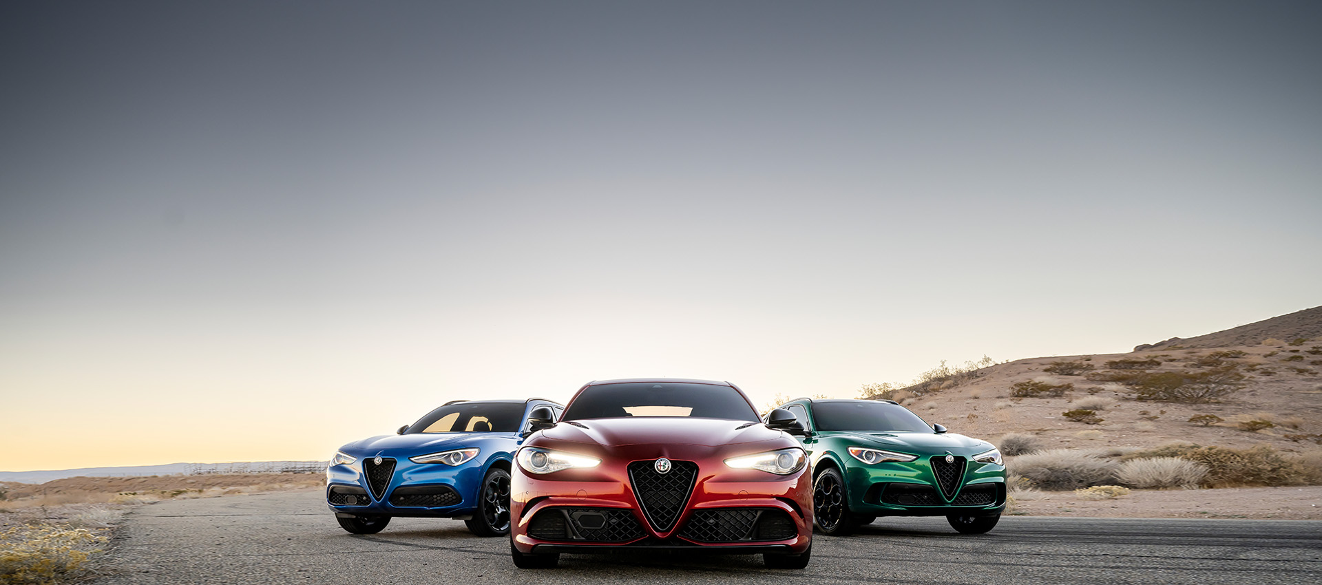Tres modelos de Alfa Romeo 2023 estacionados junto a una colina. De izquierda a derecha: un Alfa Romeo Stelvio Quadrifoglio 2023 azul, un Alfa Romeo Giulia Quadrifoglio 2023 y un Alfa Romeo Stelvio Quadrifoglio 2023 verde.