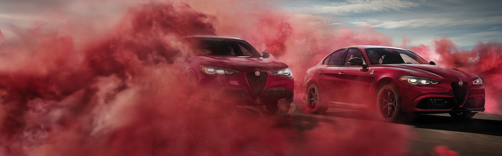 Un Alfa Romeo Stelvio Quadrifoglio 2024 rojo al lado de un Alfa Romeo Giulia Quadrifoglio 2024 rojo, en una carretera, con humo rojo envolviendo a ambos vehículos.